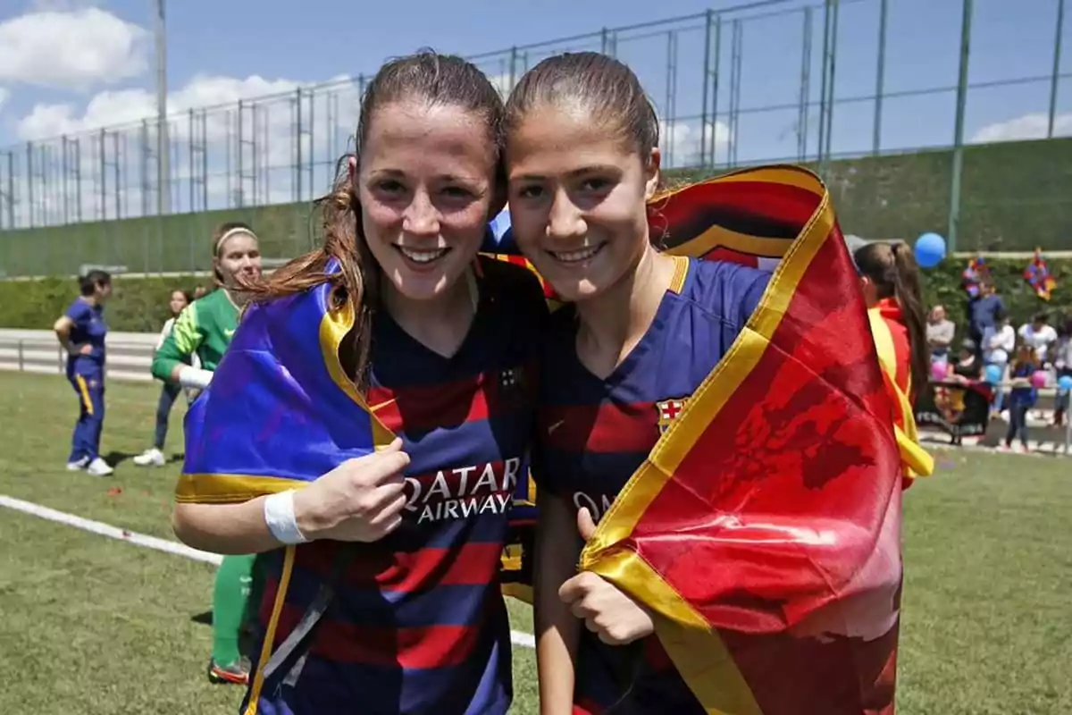 Dos jugadoras de fútbol sonríen abrazadas en el campo, llevan la camiseta del FC Barcelona y están envueltas en banderas del club Dos jugadoras de fútbol sonríen abrazadas en el campo, llevan la camiseta del FC Barcelona y están envueltas en banderas del club