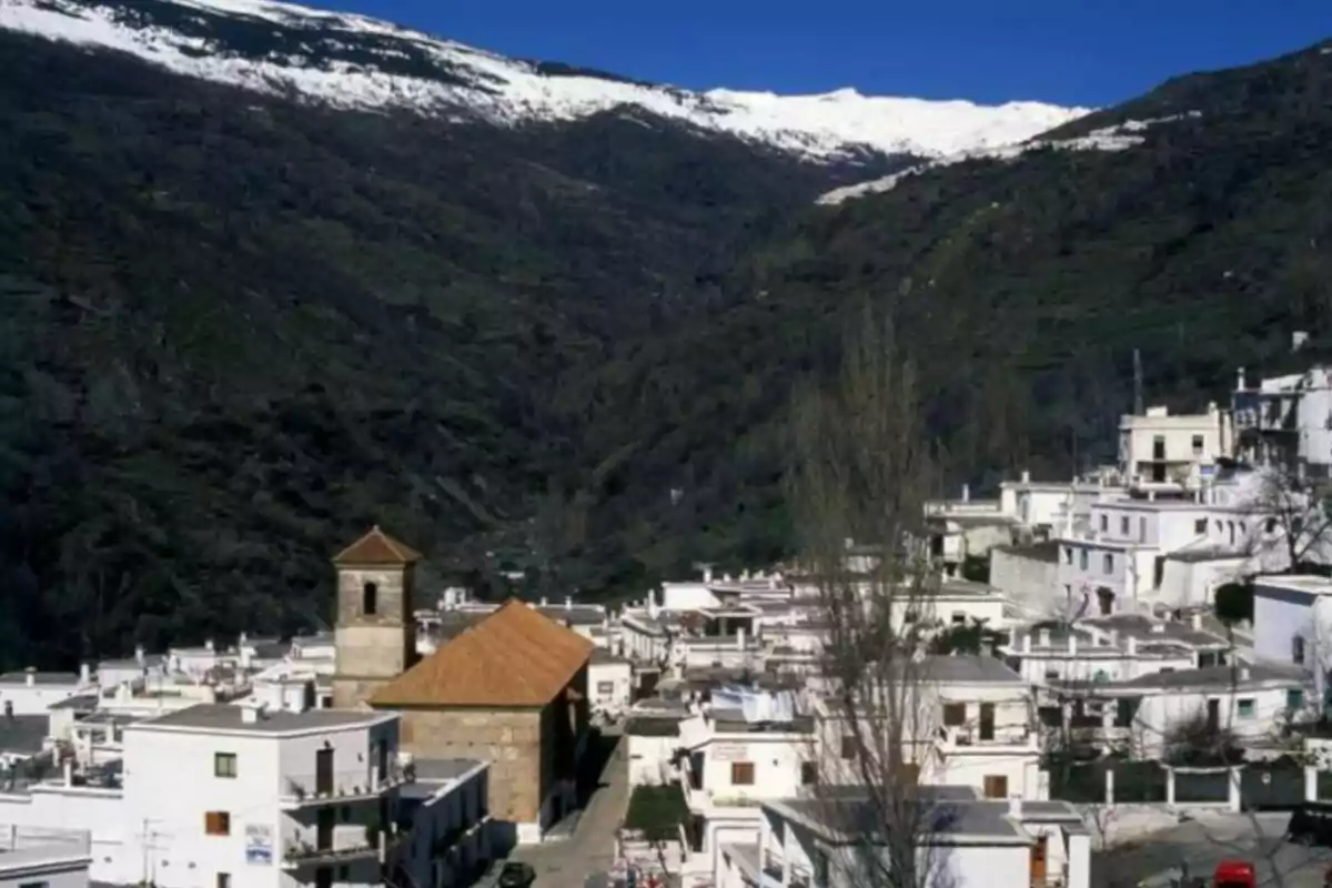 Pueblo blanco con casas de tejados planos y una iglesia con campanario, rodeado de montañas con nieve en la cima.