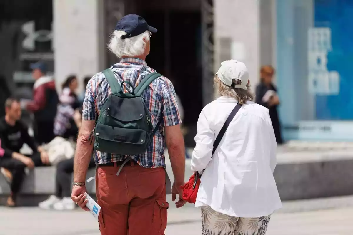 Una pareja de jubilados de espaldas, con gorra y mochila, caminando por la calle Una pareja de jubilados de espaldas, con gorra y mochila, caminando por la calle