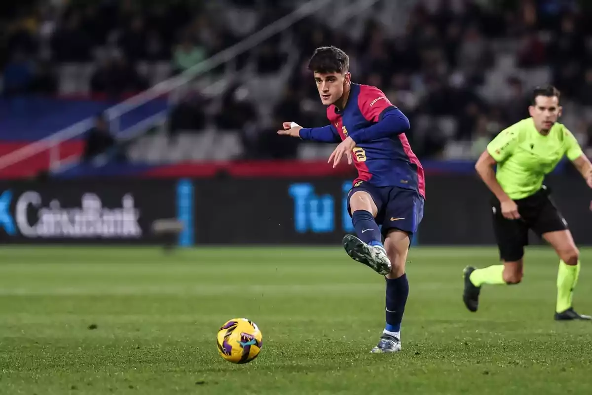 A soccer player in a blue and red uniform kicks a ball in a stadium as a referee watches. A soccer player in a blue and red uniform kicks a ball in a stadium as a referee watches.