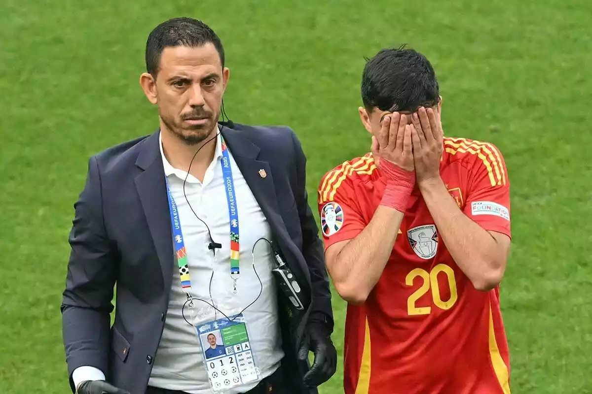 Un hombre con traje y auriculares camina junto a Pedri con uniforme rojo de España que se cubre la cara con las manos.