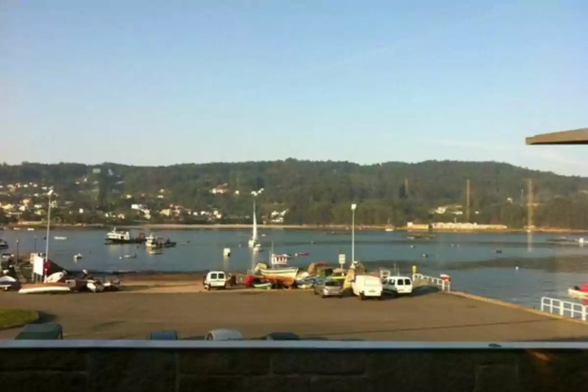 Vista de un puerto con barcos y vehículos estacionados, con colinas y casas al fondo bajo un cielo despejado. Vista de un puerto con barcos y vehículos estacionados, con colinas y casas al fondo bajo un cielo despejado.