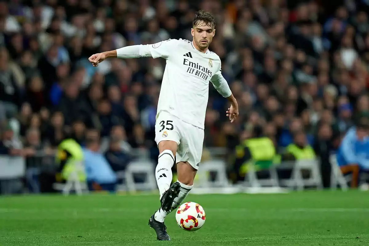 Jugador de fútbol con uniforme blanco pateando un balón en un estadio lleno de espectadores.