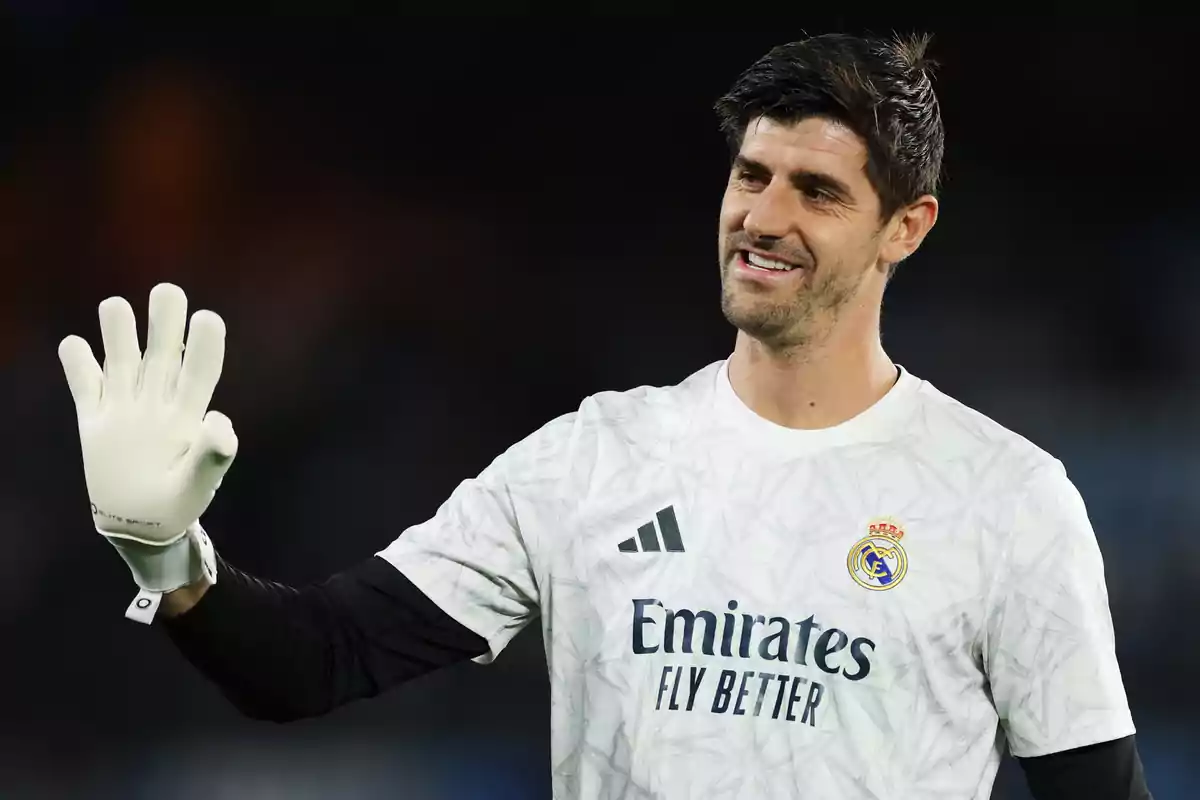 A soccer player in a white uniform and gloves raises his hand and smiles on the field.