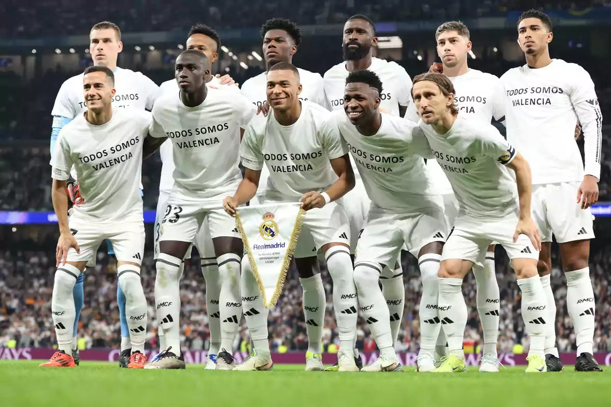 A group of soccer players wearing white T-jerseys reading "We are all Valencia" pose together in a stadium holding a Real Madrid banner.