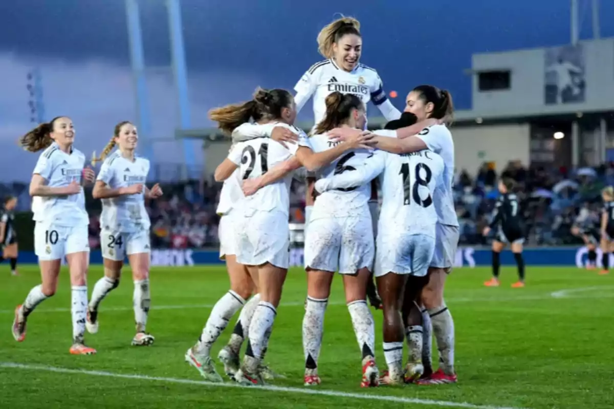 Jugadoras de fútbol celebrando un gol en el campo, vistiendo uniformes blancos y abrazándose con alegría.