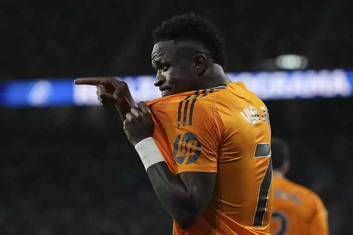 Vinicius con camiseta naranja señala con el dedo mientras sostiene su camiseta del Real Madrid.