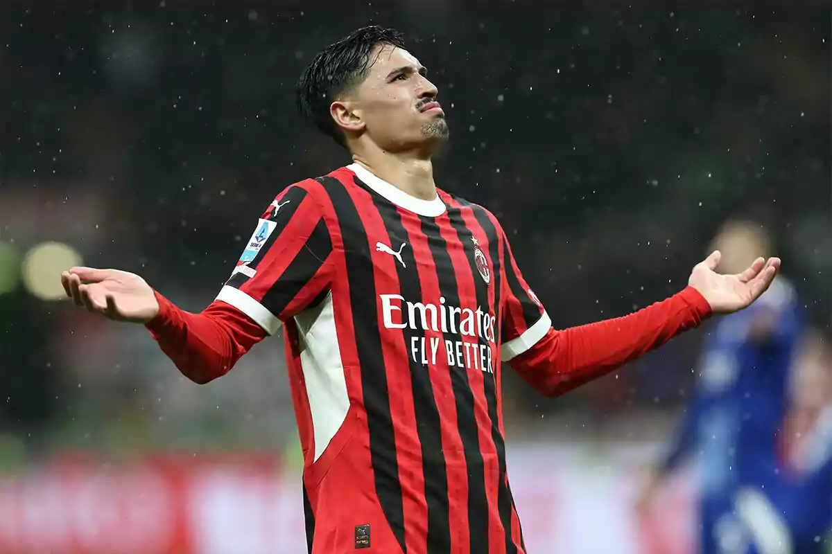 A player in an AC Milan uniform raises his arms in a gesture of frustration under the rain during a match.