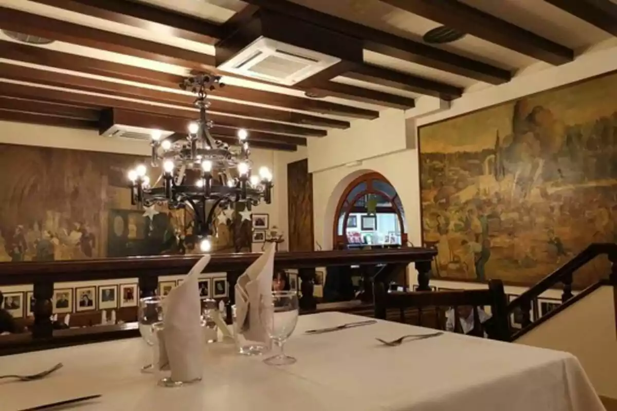 Interior de un restaurante elegante con techo de vigas de madera, candelabro colgante, mesas con manteles blancos y servilletas dobladas, y murales en las paredes.