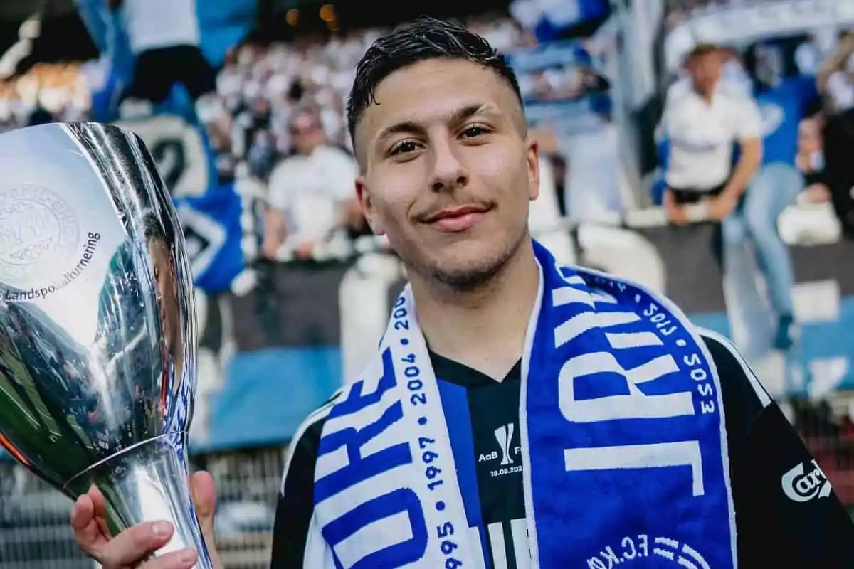Joven con bufanda azul y negra sonríe mientras sostiene un trofeo plateado en un estadio lleno de aficionados Joven con bufanda azul y negra sonríe mientras sostiene un trofeo plateado en un estadio lleno de aficionados