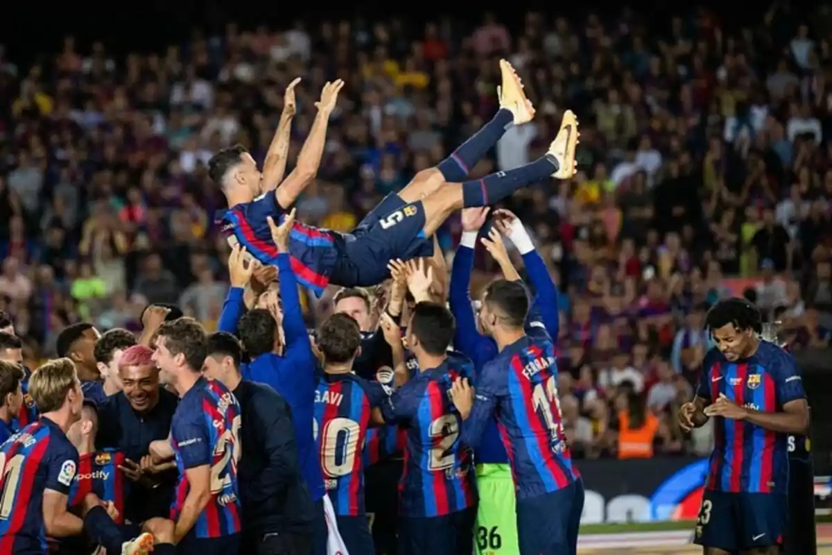 Soccer players lift a teammate in celebration during a match in a stadium full of spectators.