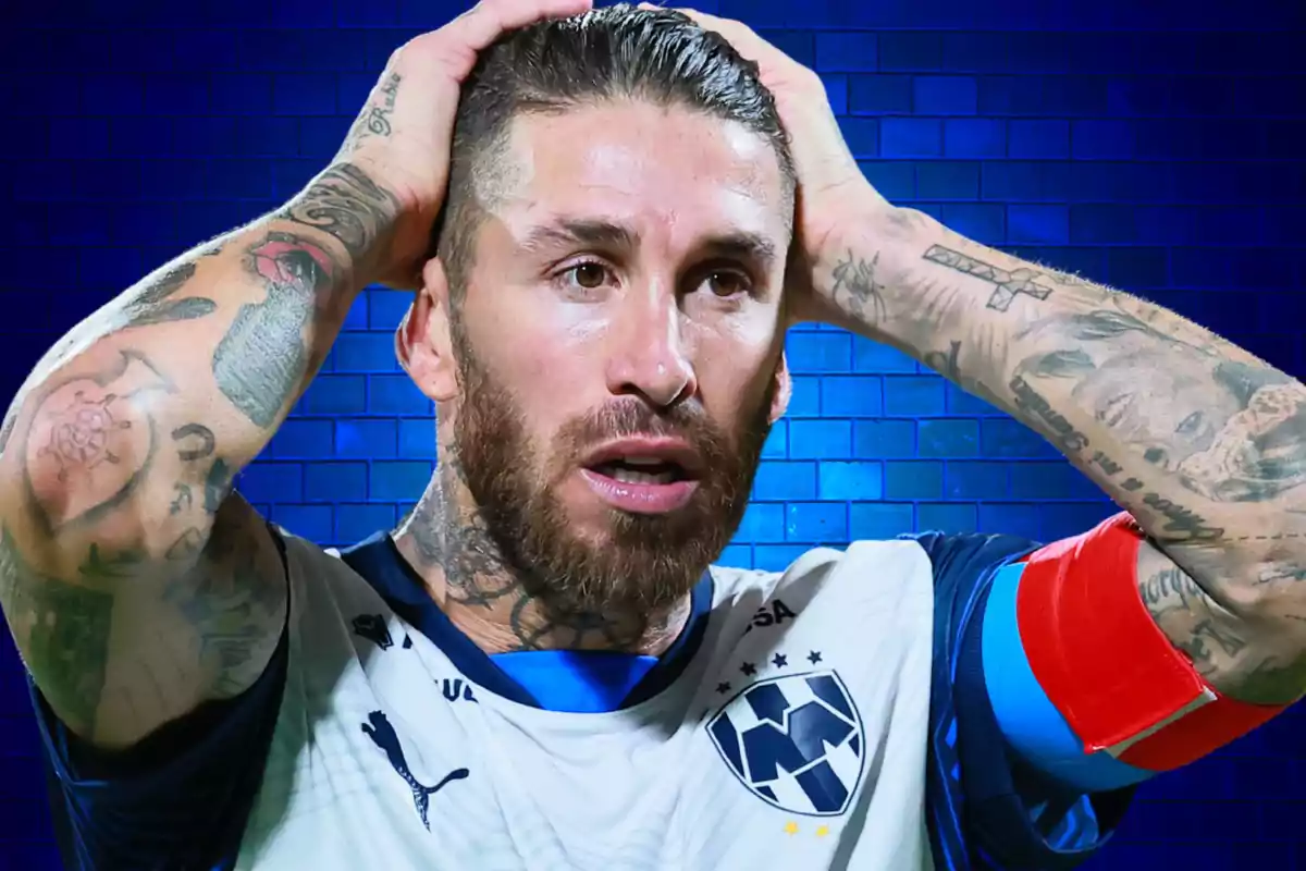 Un jugador de fútbol con tatuajes en los brazos y una camiseta blanca con detalles azules se lleva las manos a la cabeza frente a un fondo azul.