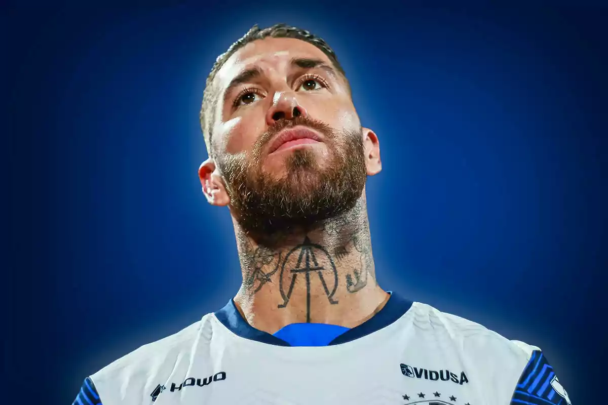 A man with a beard and neck tattoos looks up with a serious expression while wearing a white sports jersey.