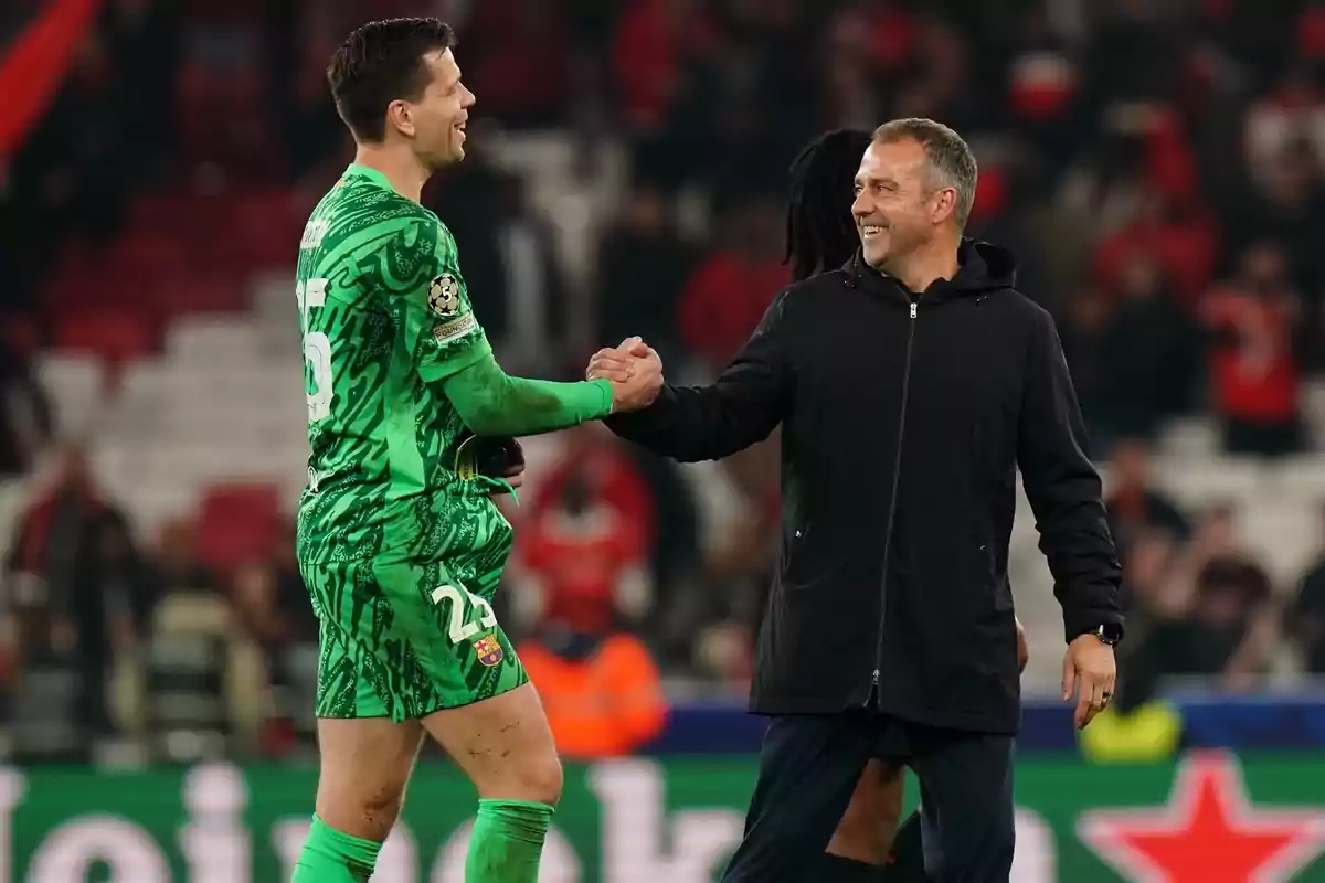 Un jugador de fútbol con uniforme verde y un hombre con chaqueta negra se dan la mano en un estadio lleno de espectadores.