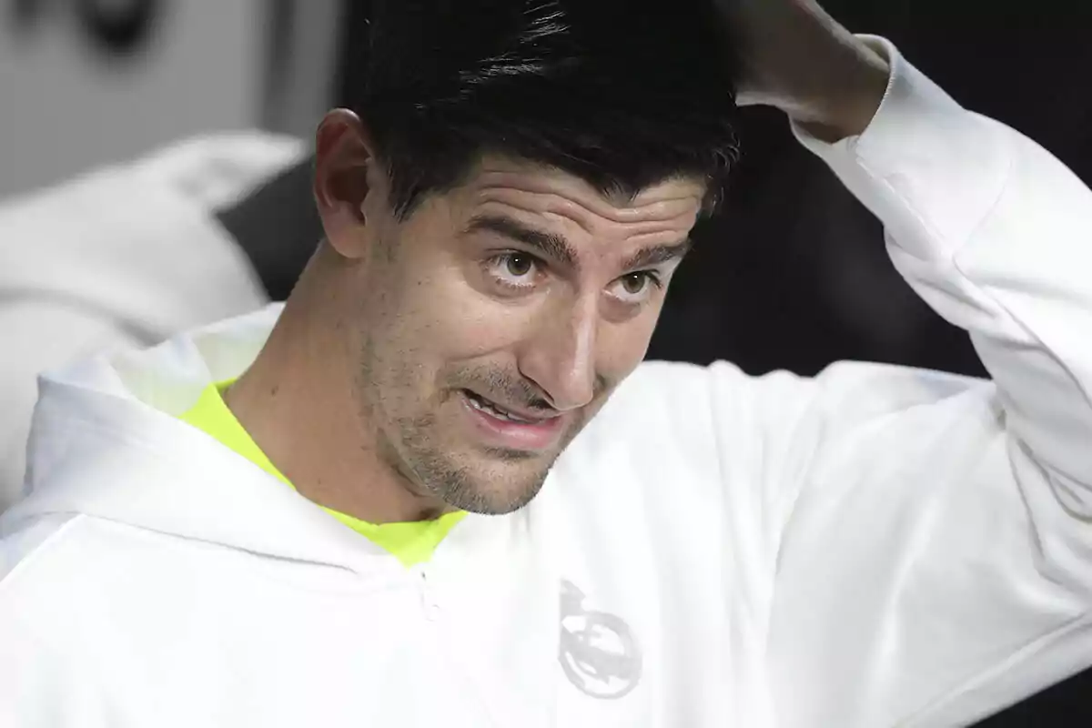 Thibaut Courtois with a thoughtful expression touches his head while wearing a white sweatjersey.