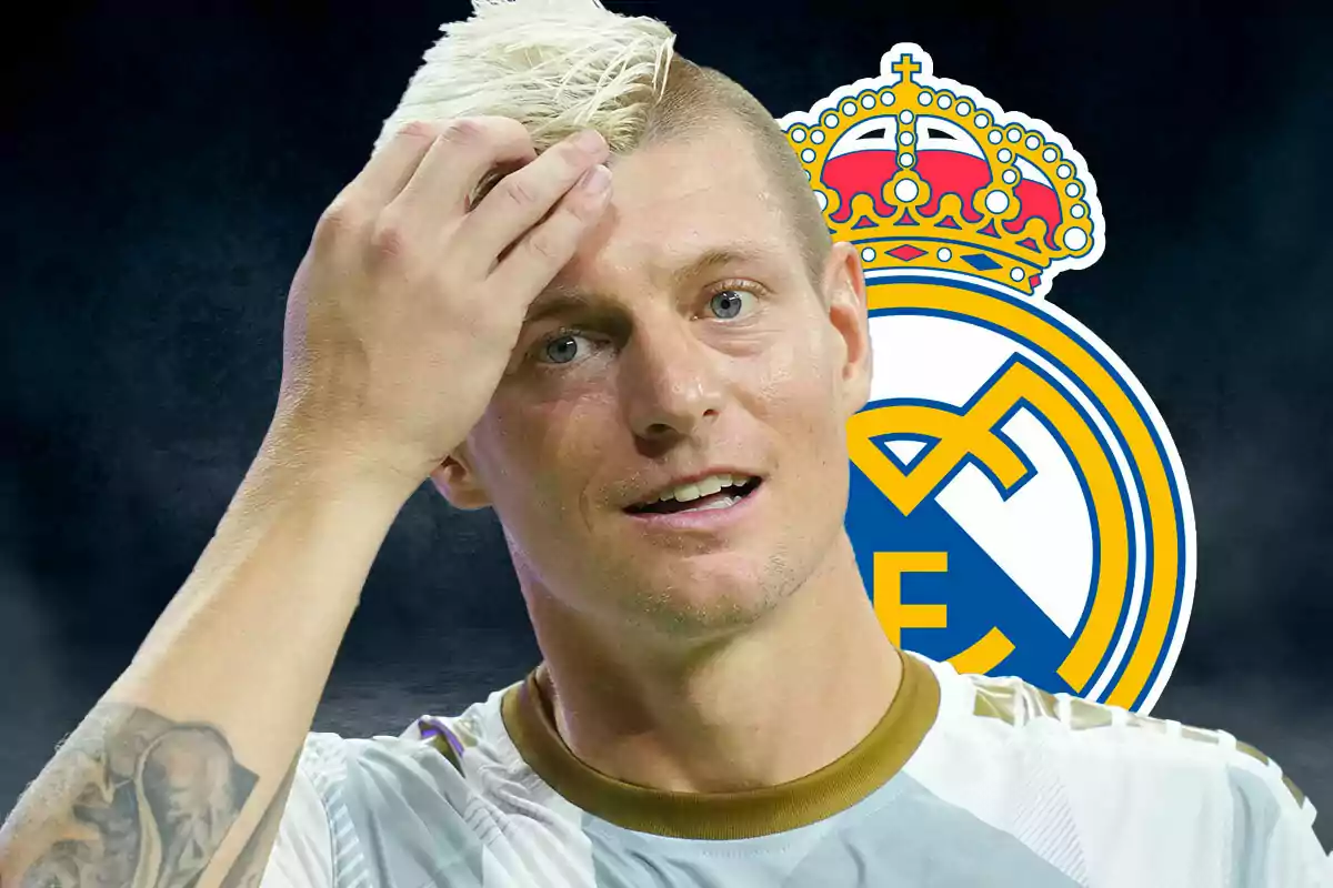 Toni Kroos, con el cabello rubio y una camiseta blanca, se toca la cabeza frente al escudo del Real Madrid.