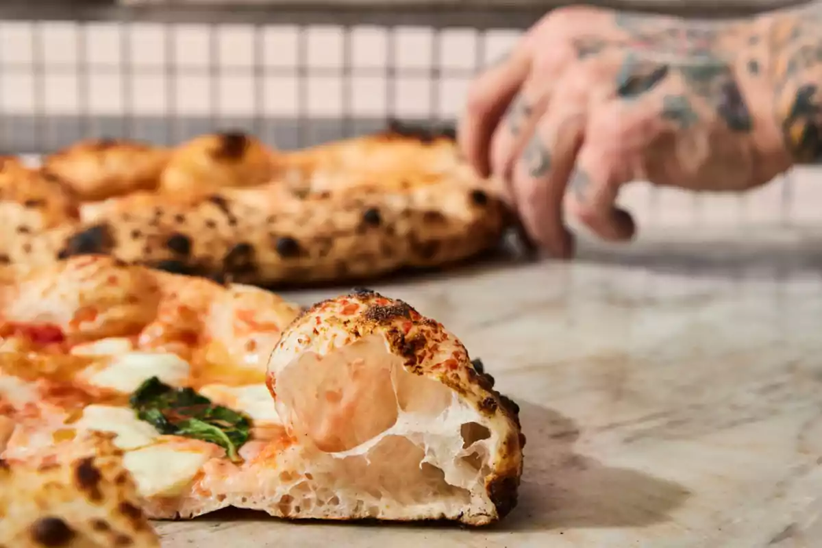 Una pizza con borde crujiente y una mano tatuada al fondo. Una pizza con borde crujiente y una mano tatuada al fondo.