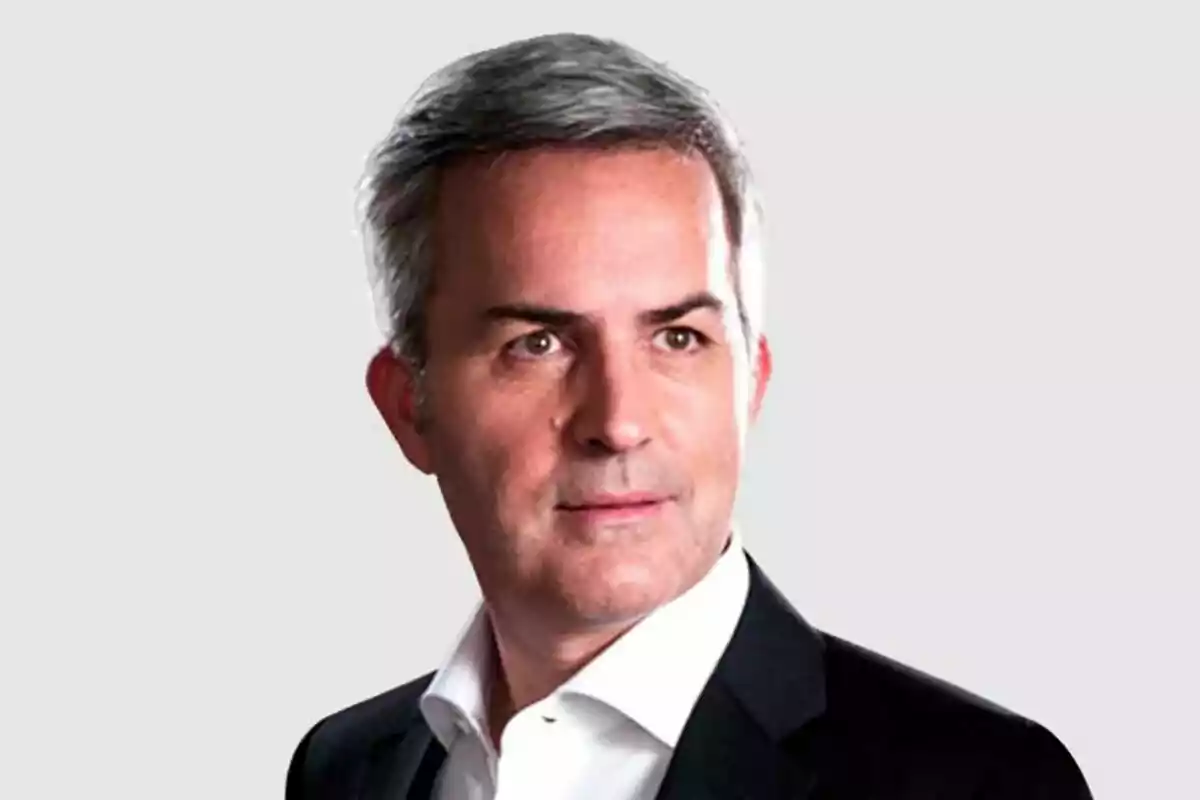 Hombre de cabello canoso con saco negro y camisa blanca sobre fondo gris claro Hombre de cabello canoso con saco negro y camisa blanca sobre fondo gris claro