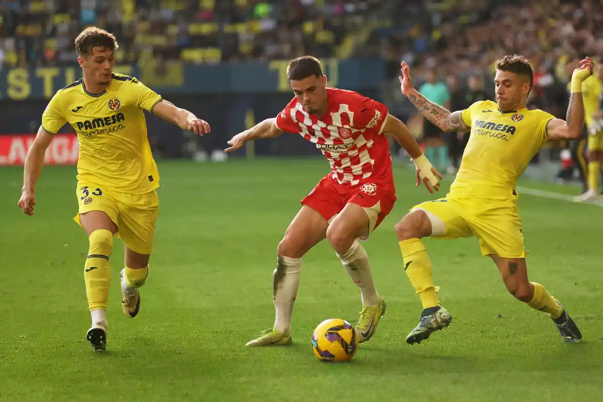 Soccer players compete for the ball in a match, one in a red and white uniform and two in yellow uniforms, in a stadium full of spectators. Soccer players compete for the ball in a match, one in a red and white uniform and two in yellow uniforms, in a stadium full of spectators.