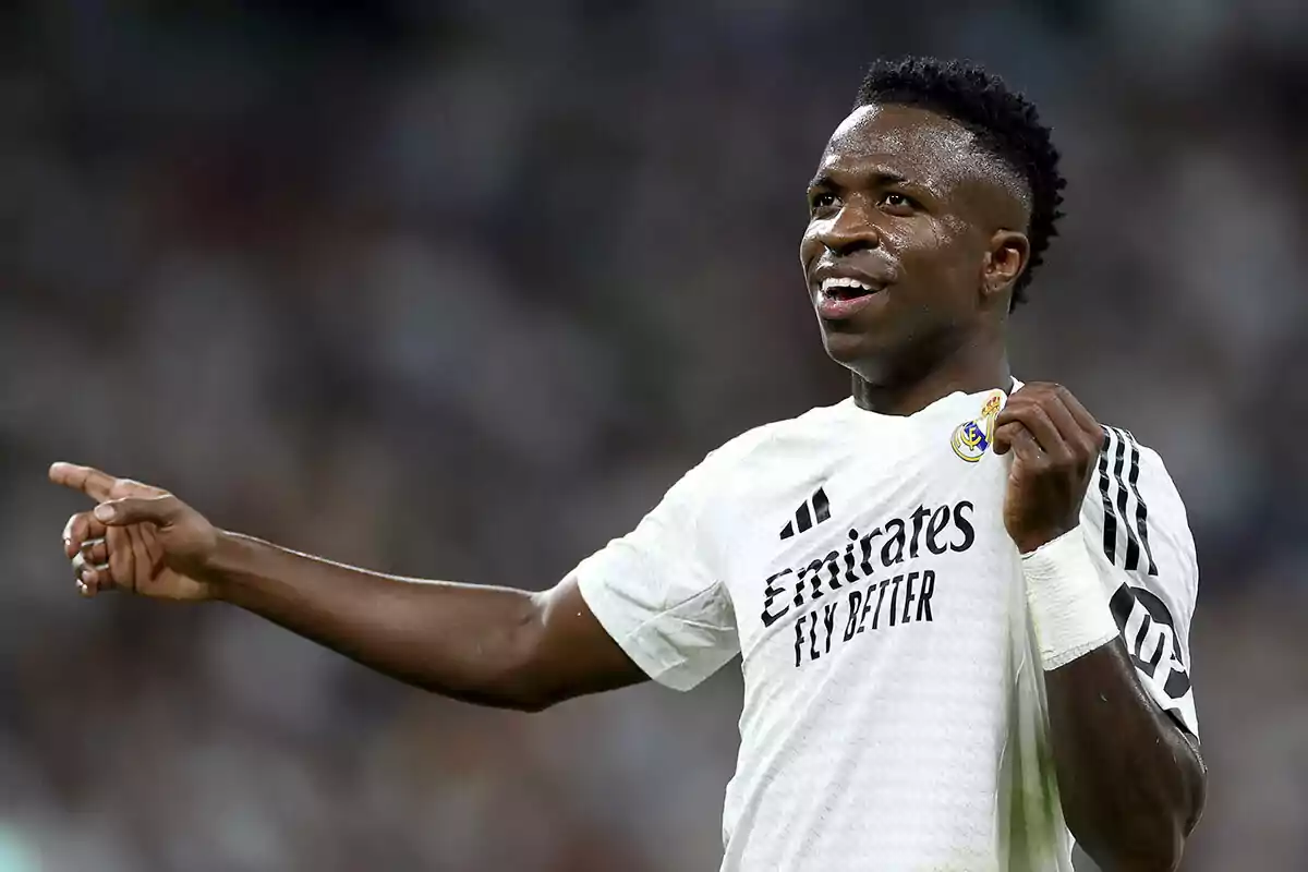 Vinicius con uniforme del Real Madrid señalando con el dedo y sosteniendo el escudo de su camiseta.