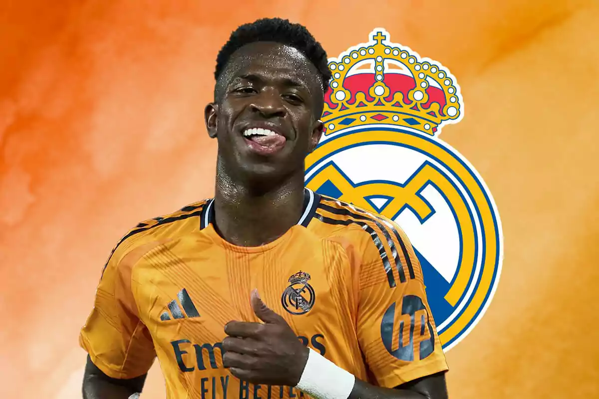 Vinicius con uniforme naranja sonríe frente al escudo del Real Madrid sobre un fondo naranja.