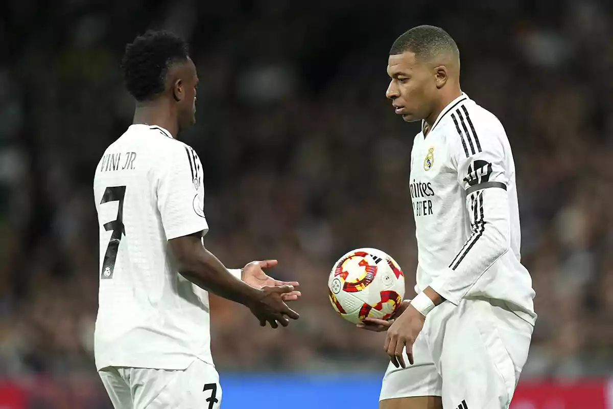 Dos futbolistas con uniforme blanco en el campo de juego, uno sosteniendo un balón.