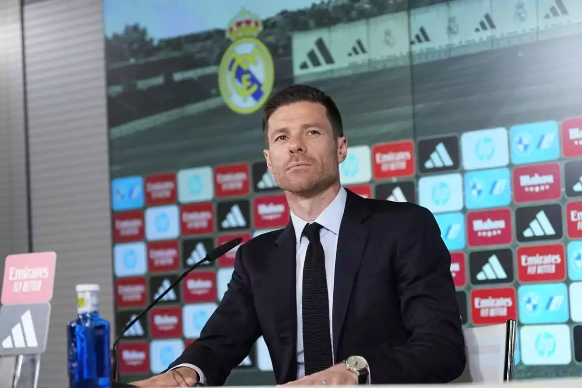 Man in a dark suit sitting at a press conference with the Real Madrid crest and a sponsor backdrop behind him