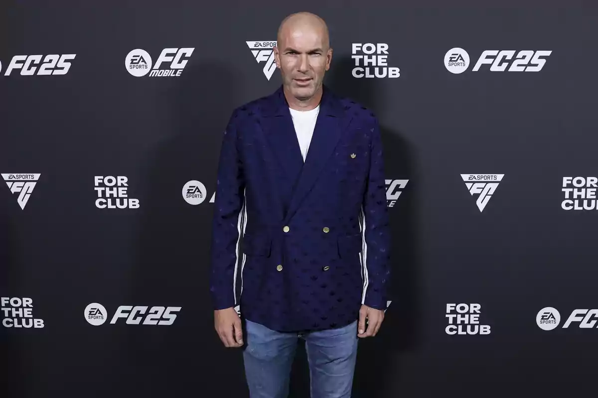 Hombre calvo con saco azul y pantalón de mezclilla posando frente a un fondo con logotipos de EA Sports FC25 y For The Club Hombre calvo con saco azul y pantalón de mezclilla posando frente a un fondo con logotipos de EA Sports FC25 y For The Club